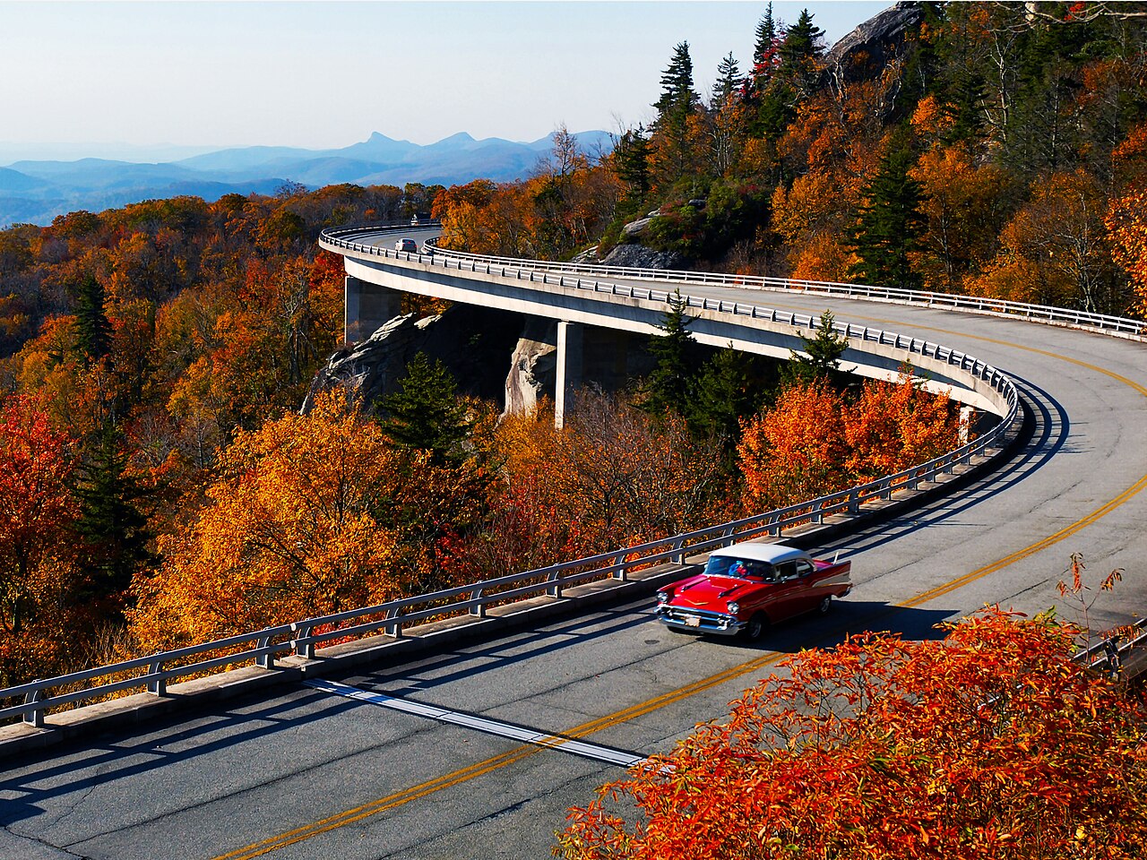 A red car on a curving road bridge that hugs a mountainside covered in fall color