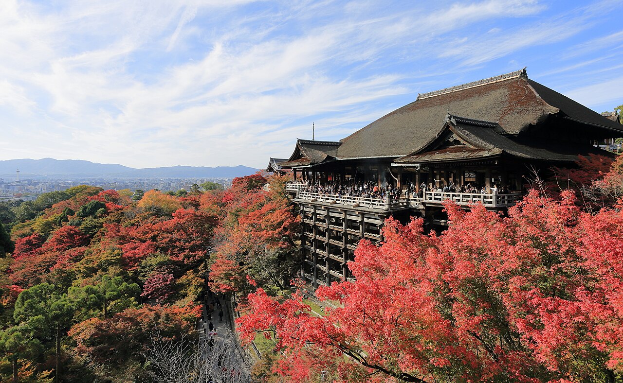 The wooden Kiyomizu-dera temple on its tall stilts above red and orange autumn trees, with Kyoto in the distance