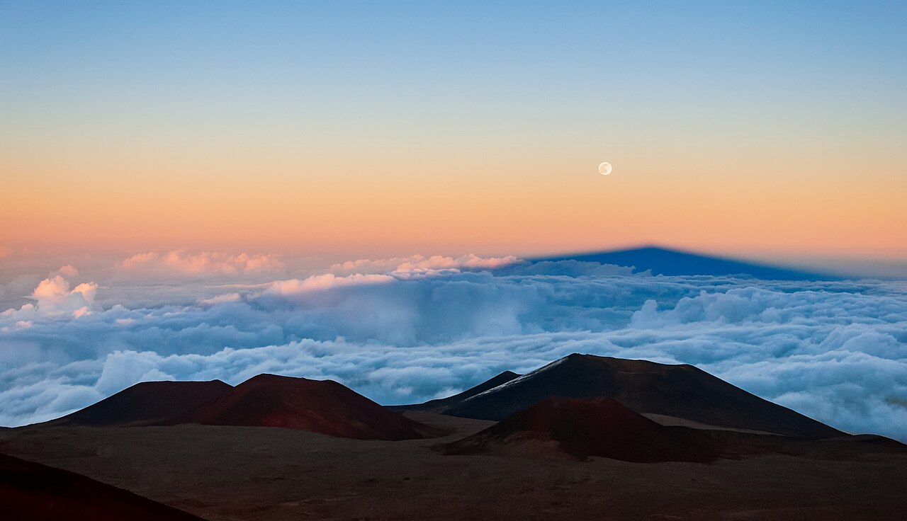 The full Moon rising in a pink and blue sky above a sea of clouds and the shadow of a mountain