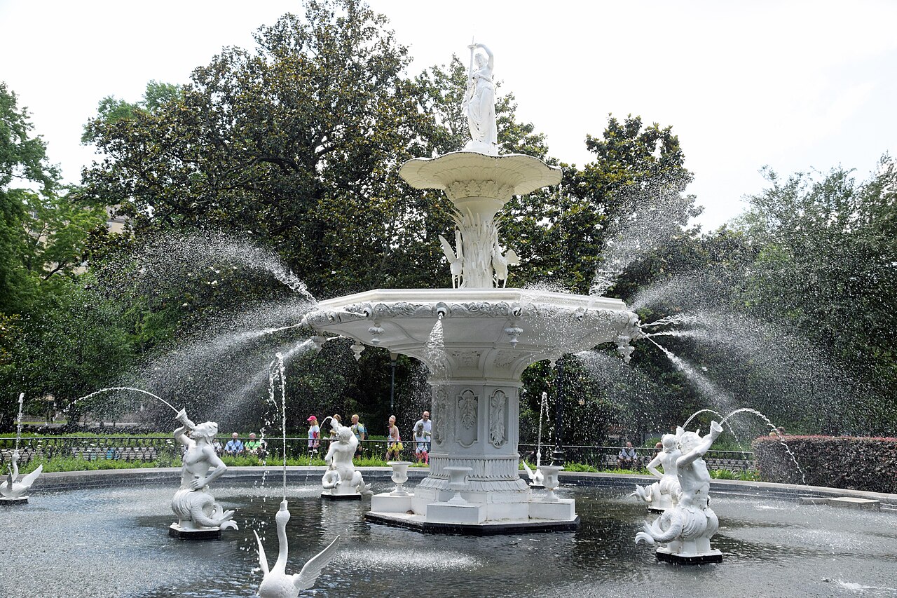 A white cast-iron fountain spraying water in a park shaded by trees