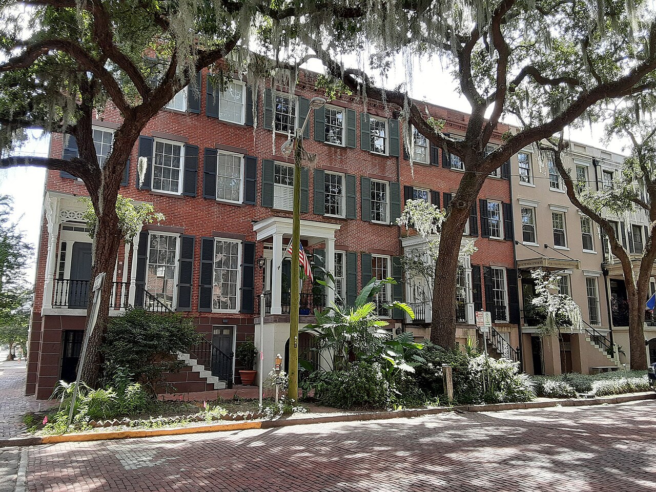 A row of red-brick townhouses with black shutters behind shady oak trees
