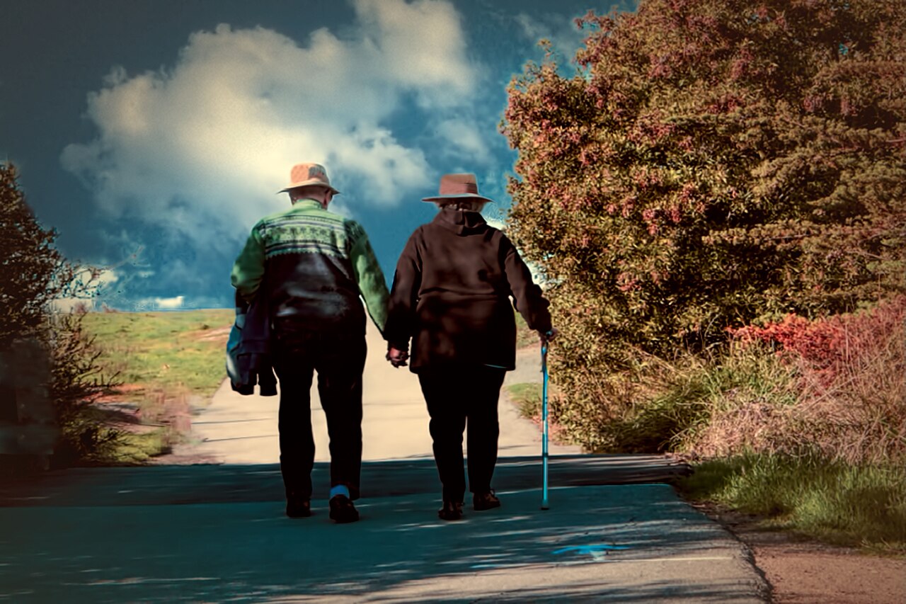 An older couple walking hand in hand along a park path, one with a walking stick